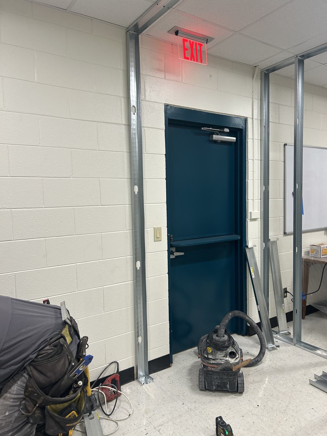 Metal stud framing for a new office wall beside a lit exit door in a school corridor, shop vacuum in place.