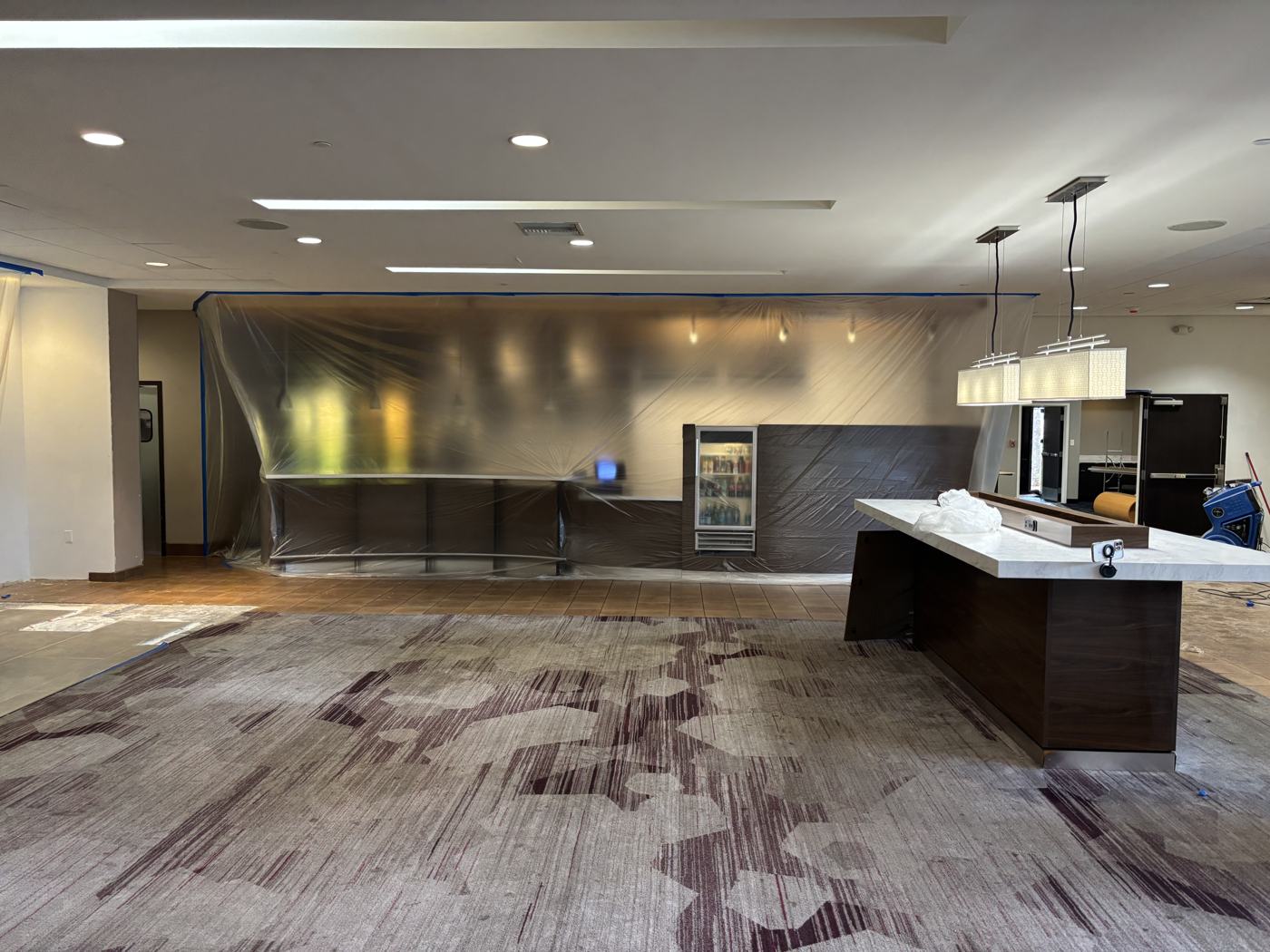 Hotel lobby bar fully wrapped in protective sheeting, taped at the ceiling line, with the lobby carpet and pendant lights intact.